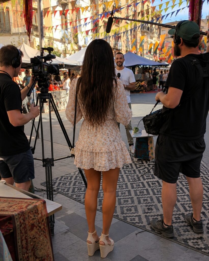 Television production crew filming a presenter at a medieval market in Alicante surrounded by artisan stalls and colourful festival decorations.