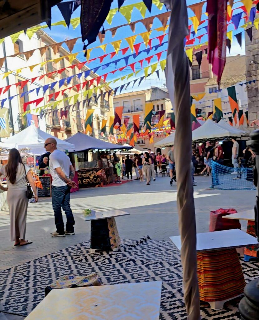 Colourful medieval market square in Alicante filled with artisan stalls, historic buildings, festive bunting and visitors enjoying the event.